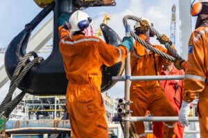 Offshore workers attaching a large sling to a crane block for heavy lifting from a construction barge to an oil platform.