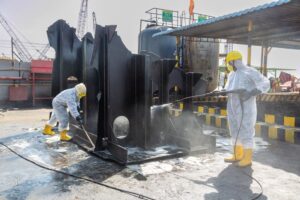 Workers in protective suits and yellow boots cleaning oily ship parts at an Indian ship recycling yard, showcasing industrial safety measures.