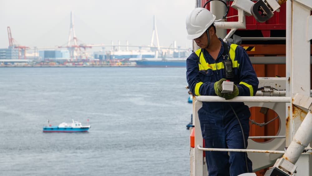 A seaman crew member aboard a cargo vessel, equipped with personal protective equipment, operates the lifeboat davit while wearing a helmet and using a walkie-talkie.