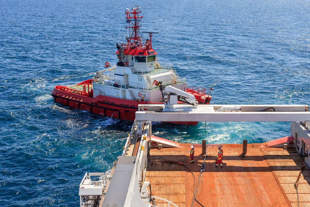 The deck crew is engaged in offshore work, with a small tugboat positioned behind a larger Anchor Handling Tug Supply (AHTS) vessel.






