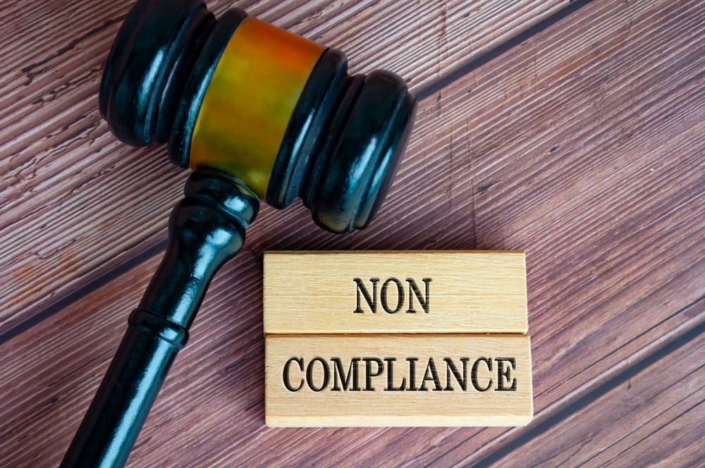 The image featuring the wooden blocks engraved with "Non Compliance" and the lawyer's gavel in the background, emphasizing legal consequences.
