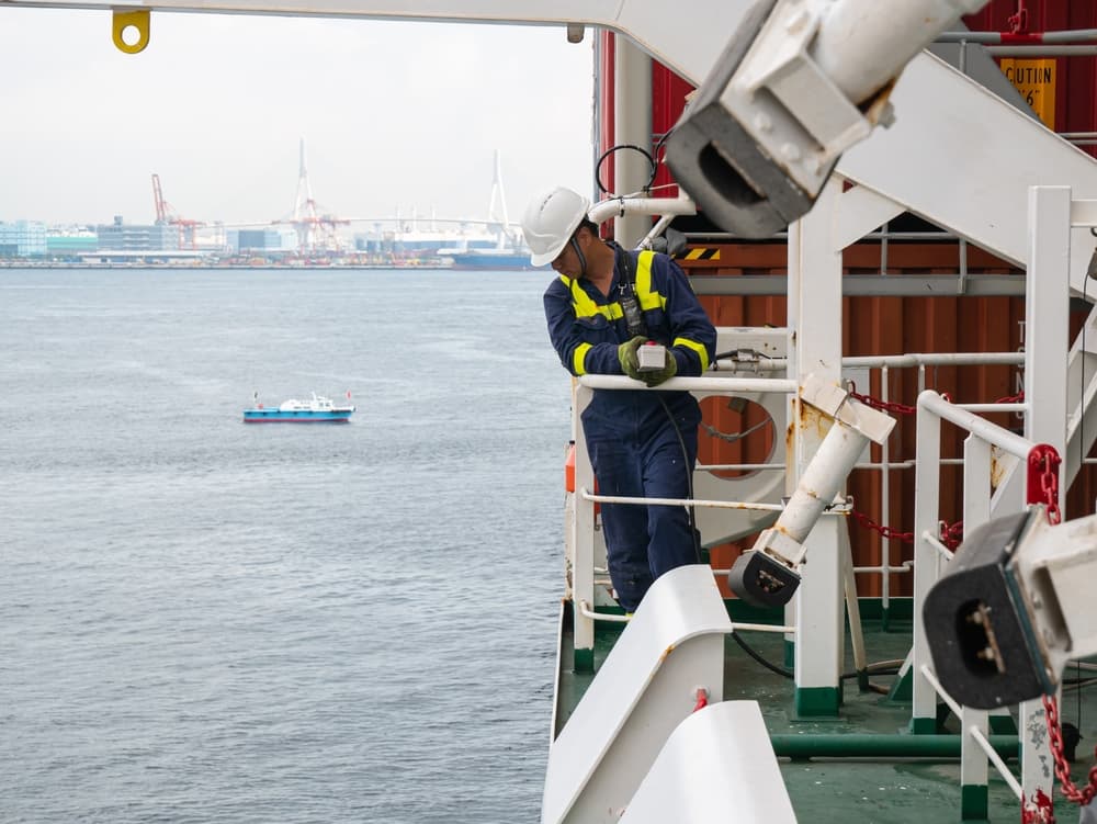 Cargo ship crew member in helmet and PPE operating lifeboat davit while using a walkie-talkie.