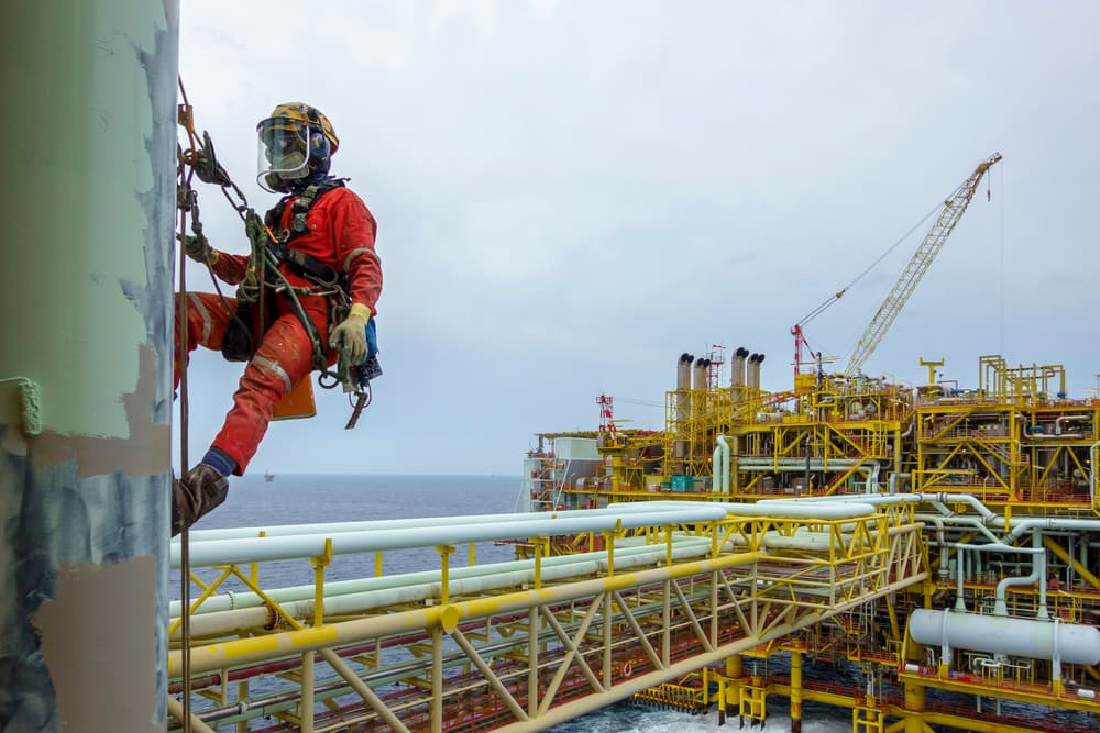 A worker performing duties at height, equipped with Personal Protective Equipment (PPE) and a fall protection system, suspended from a pipeline for painting tasks.






